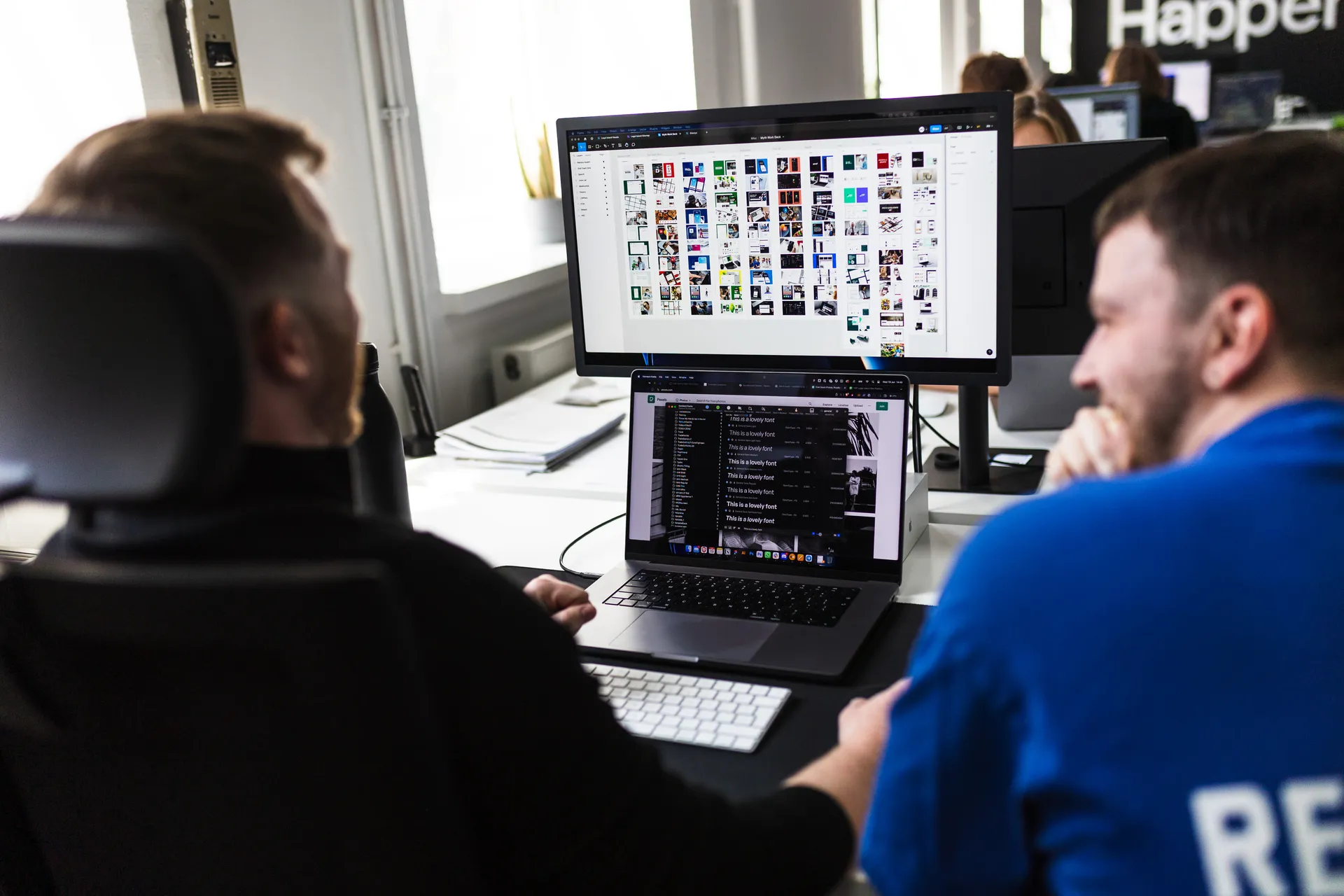 Two developers with a monitor full of design thumbnails and code on a laptop